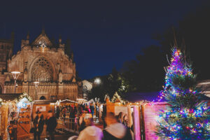 Busy crowds of people visiting the twinkling festive chalets at Exeter Cathedral Christmas Market. The west front of Exeter Cathedral is in the background and a christmas tree is present in the foreground.