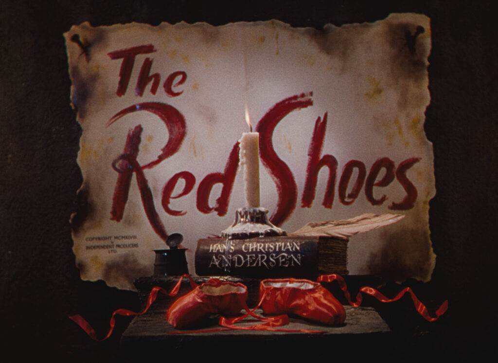 Behind a candle on a book is burned paper with the title The Red Shoes, in front of the candle are red ballet shoes