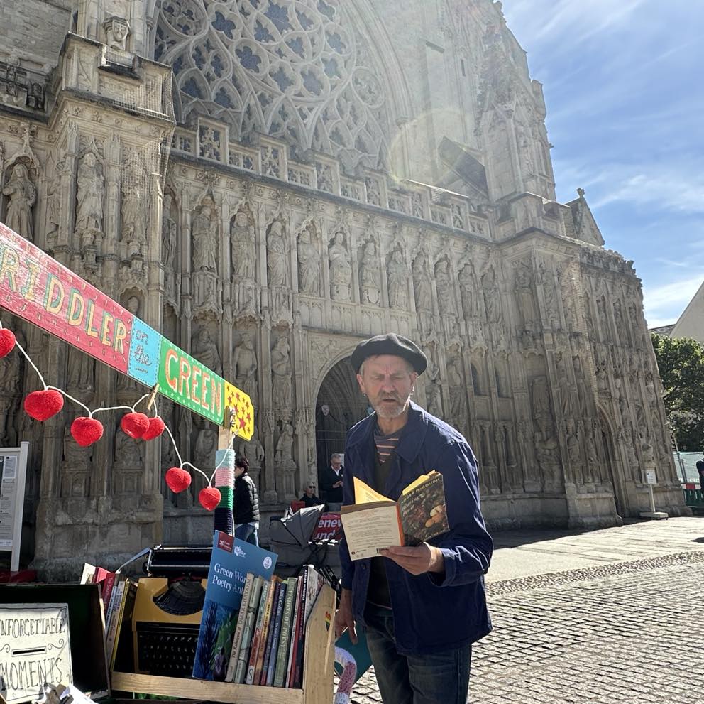 Exeter Cathedral's Riddler in Residence, Jon Seagrave aka Jonny Fluffypunk, standing on the west front of Exeter Cathedral reading a book next to his poetry pram - a decorated pram filled with poetry books and typewriter.